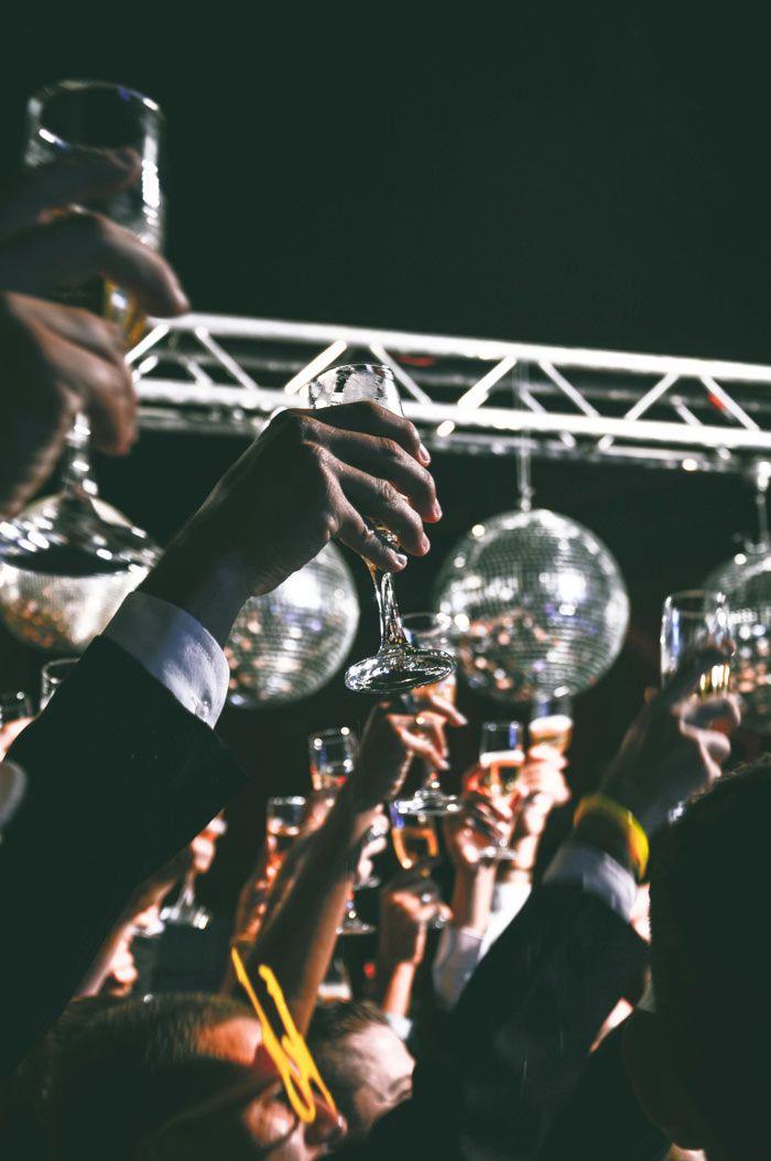 A crowd raises glasses in a toast under disco balls and stage lights at a lively party.